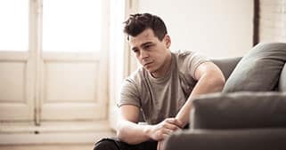 A young man in a gray t-shirt sits on the floor next to a couch, looking sad and thoughtful in a softly lit room with large windows in the background.
