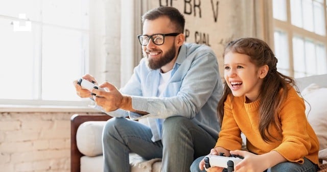 A smiling man and young girl sit on a couch, playing video games together with controllers in a bright, cozy living room—a perfect moment that highlights the positive impact of a co-parenting program for dads.