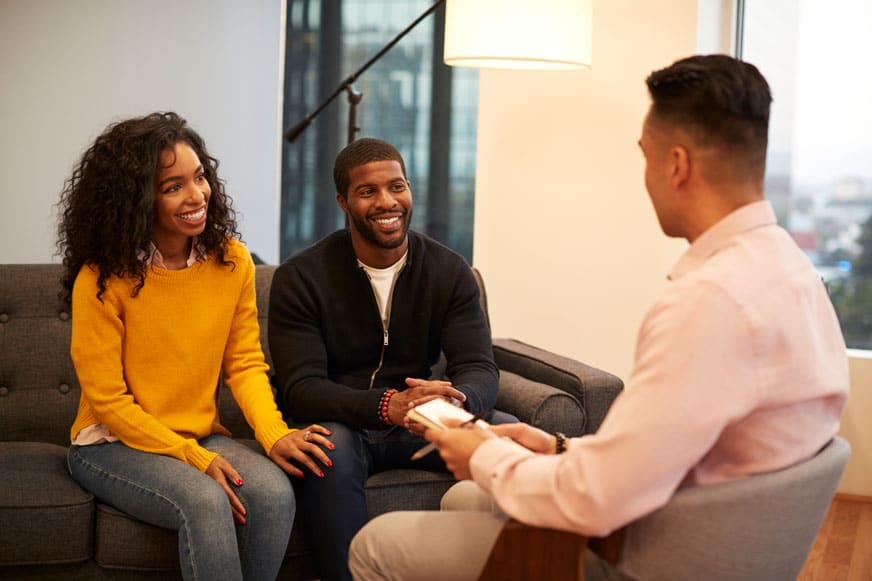 A smiling couple sits on a couch, facing a person holding a clipboard and pen, suggesting a meeting or counseling session in a modern office setting.
