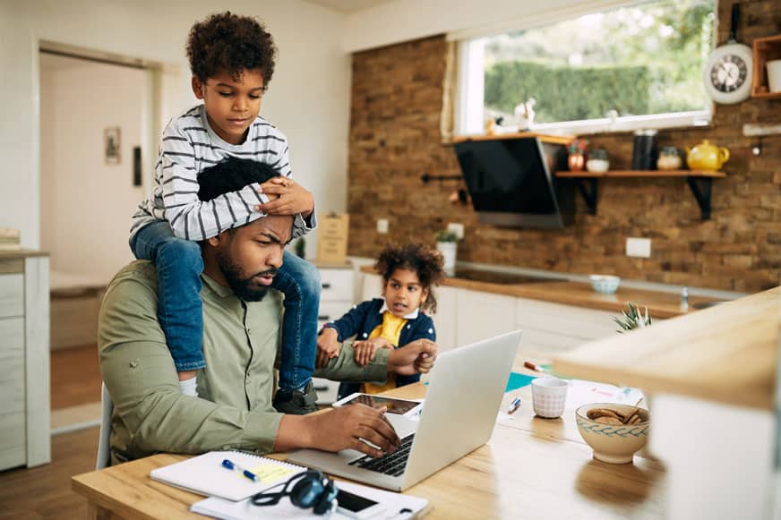 A man works on a laptop at a kitchen table while a child sits on his shoulders and another child draws beside him. Papers, headphones, and a bowl are on the table.
